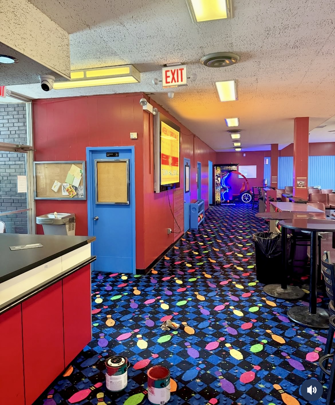 A hallway with colorful bowling pin carpet, red walls, and fluorescent lighting, showing an arcade game in the distance.