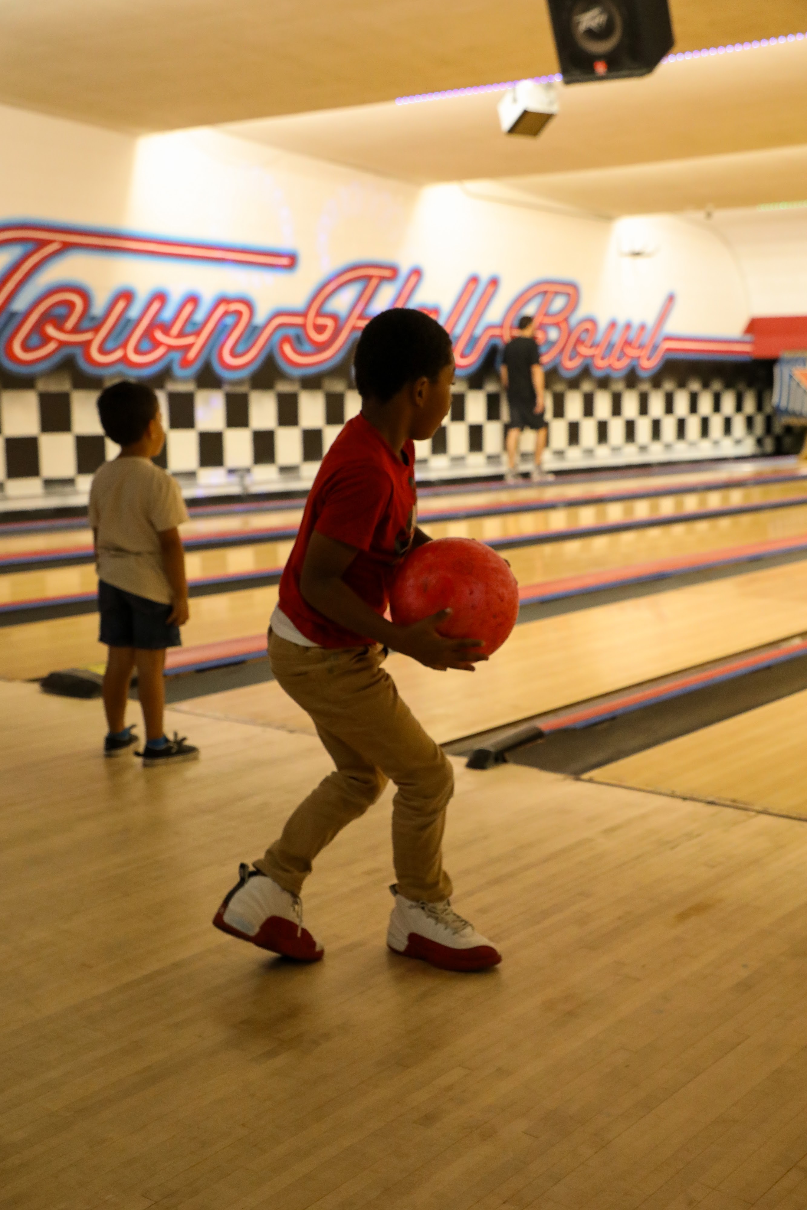 A young boy in a red shirt holds a red bowling ball on a lane, preparing to bowl at Town Hall Bowl.