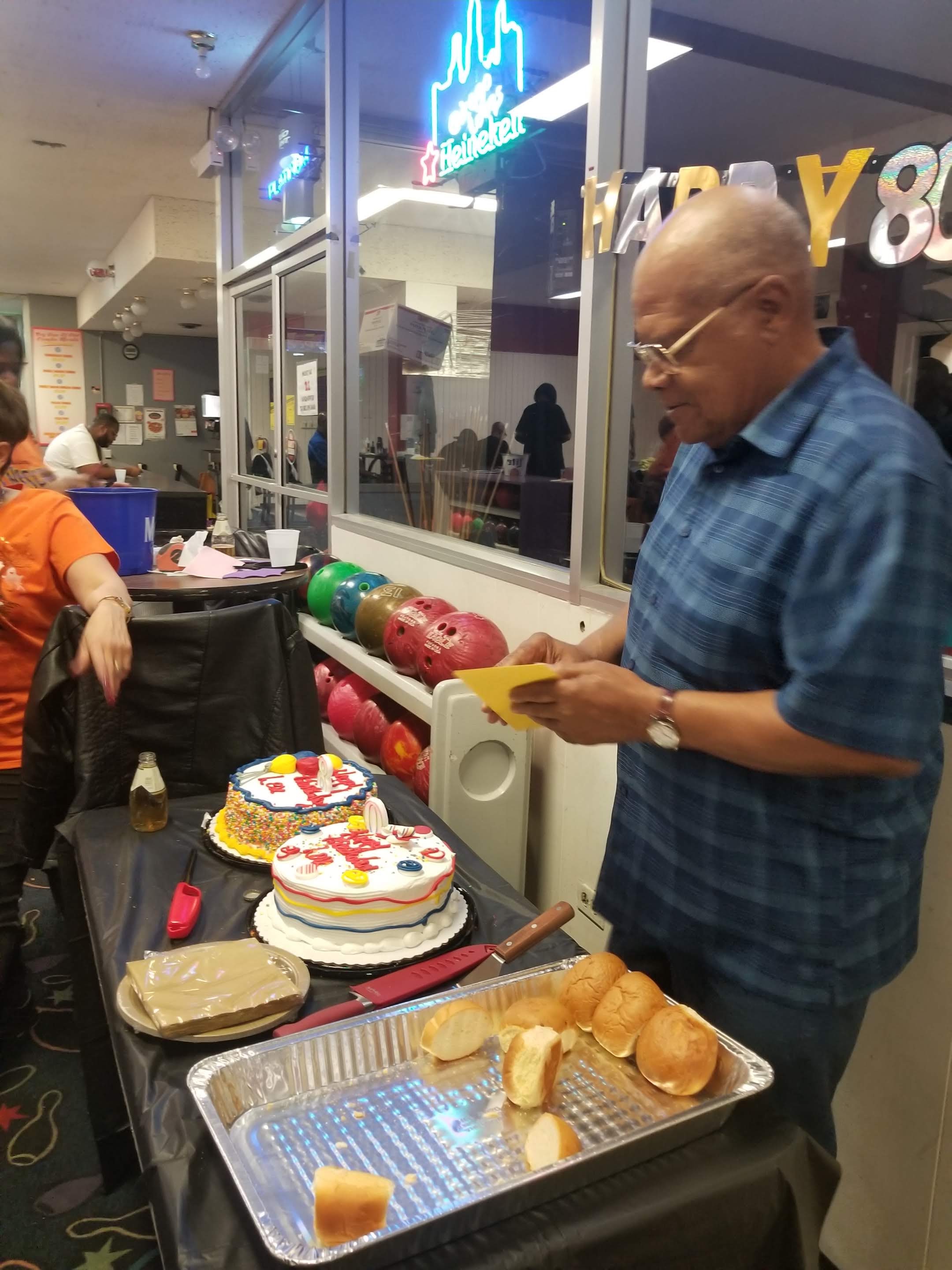 A man holds a card at an 80th birthday party with cakes and bowling balls visible at a bowling alley.