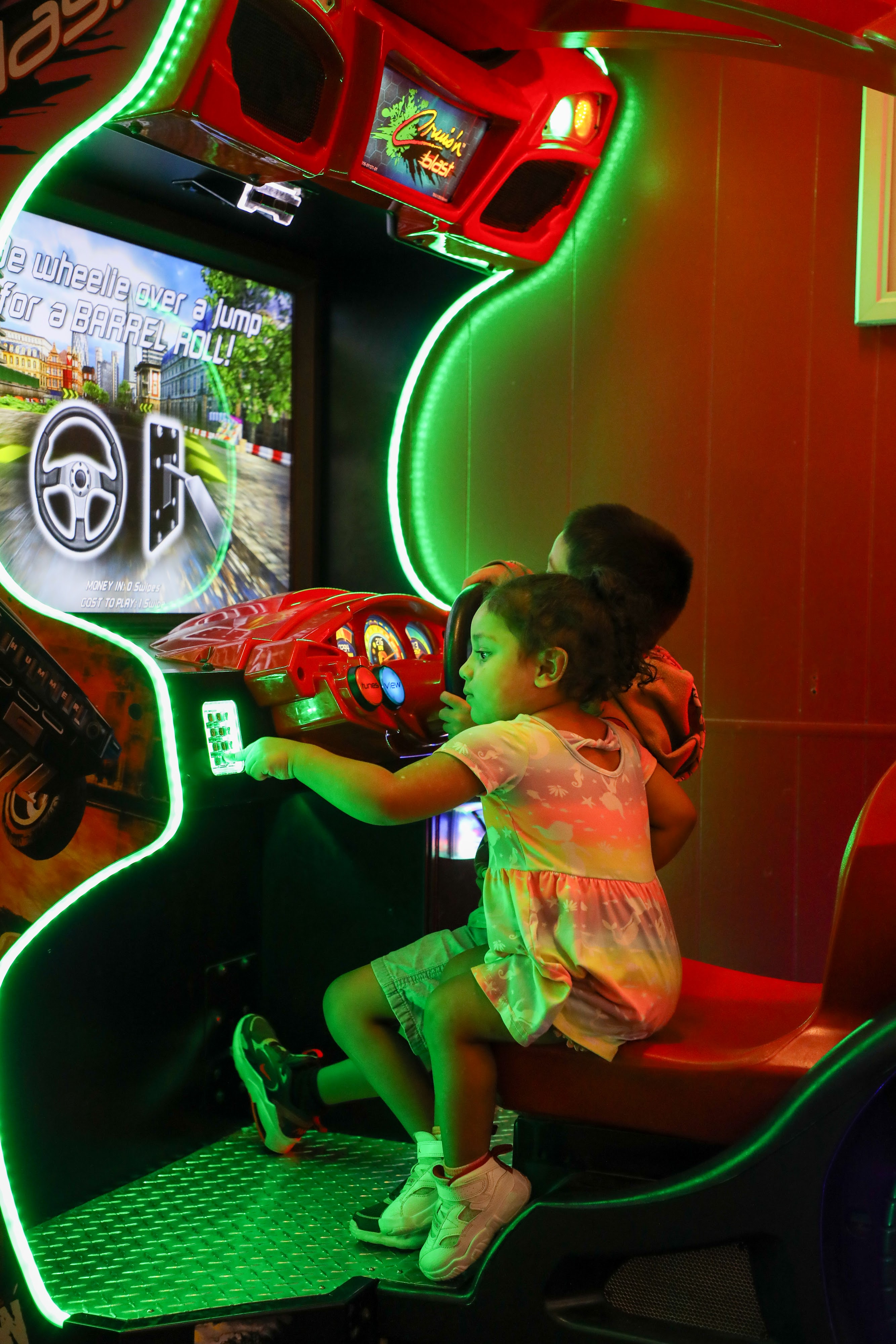A child in a tie-dye dress plays a 'Cruis'n Blast' racing arcade game, with bright green neon lights illuminating the scene.