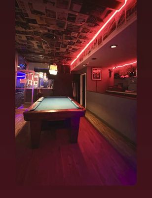 A dimly lit room with a billiard table, red neon lights, and a ceiling covered in newspaper clippings.