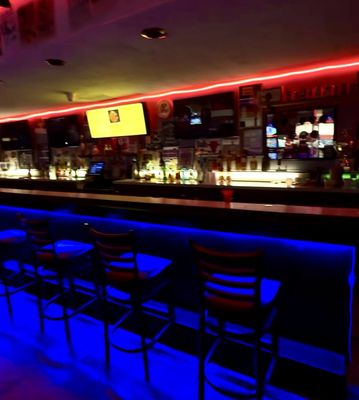 A vibrant bar area with blue under-bar lighting, red neon accents, and multiple TV screens behind the counter.