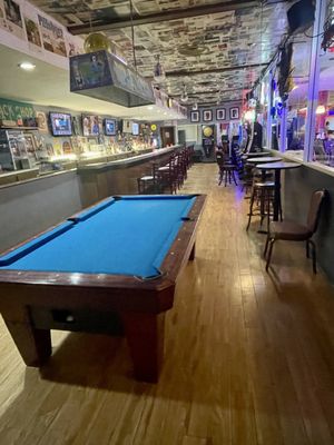 A pool table with a blue felt surface is in the foreground of a bar and lounge area.