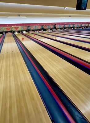 Several empty bowling lanes with red and black striped gutters, a few pins standing, and two red bowling balls.