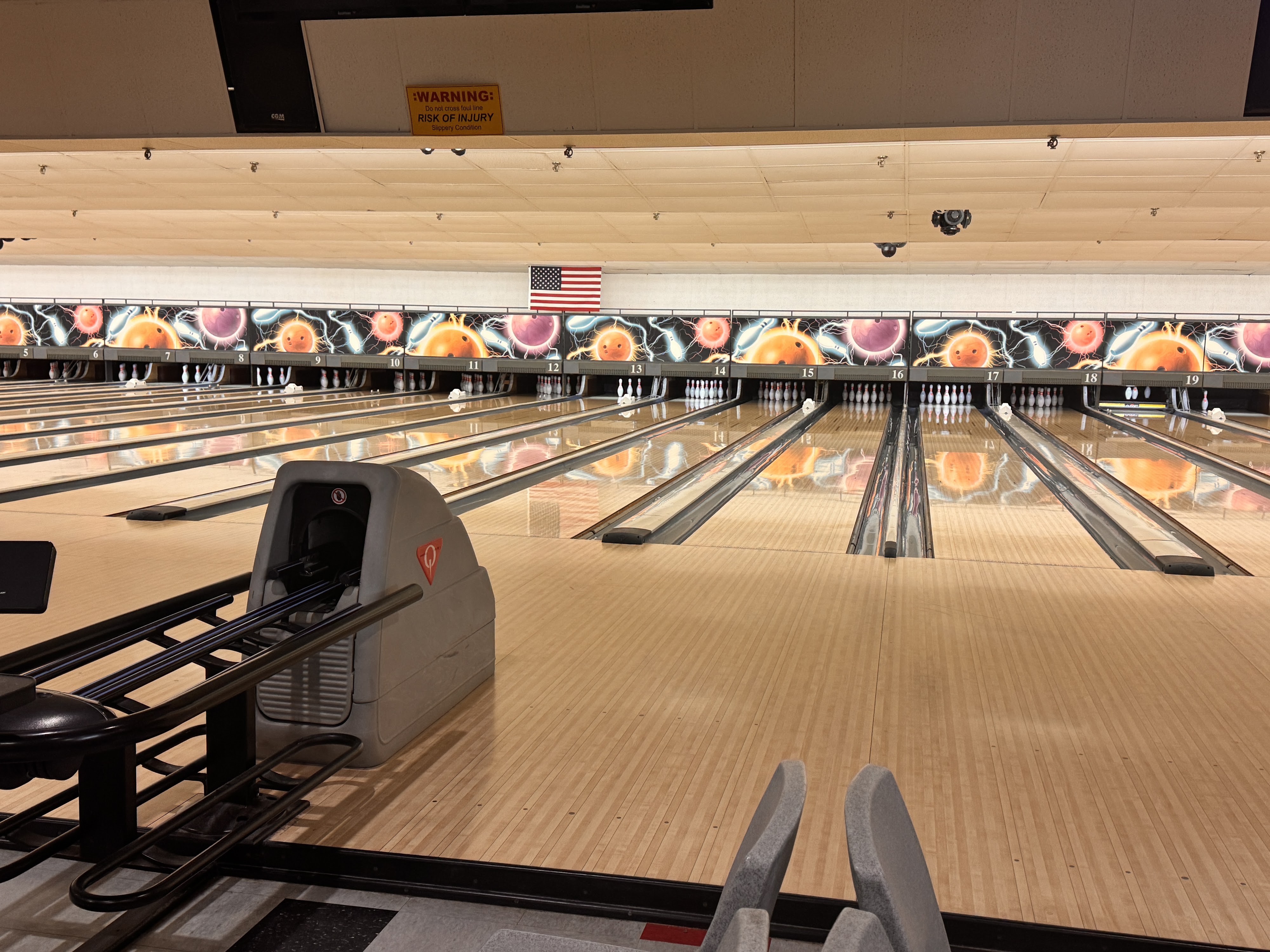 A bowling alley with multiple polished wooden lanes, vibrant retro pin setter graphics, and an American flag above the lanes.