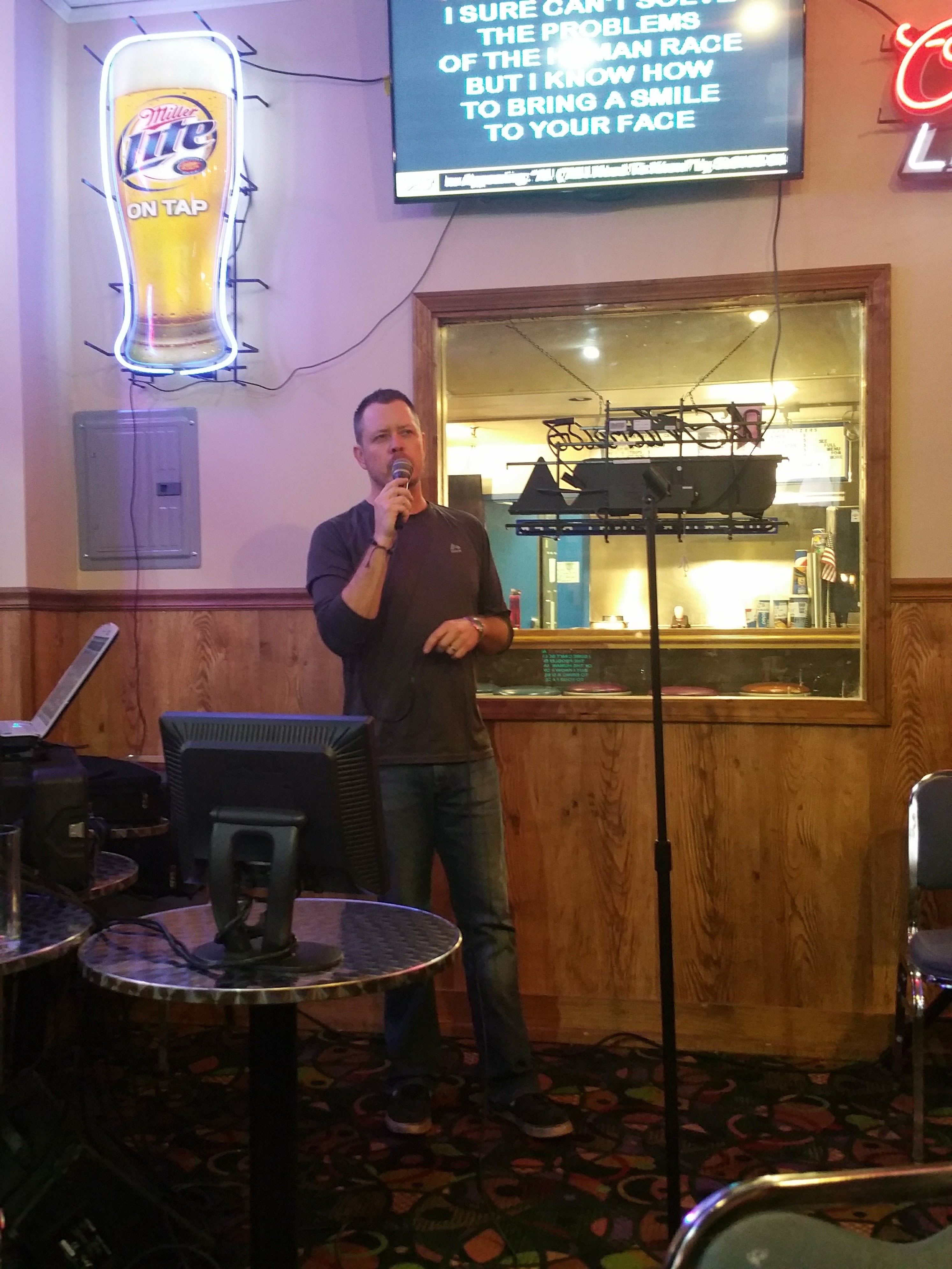 A man sings karaoke into a microphone with lyrics displayed on a screen above him and a Miller Lite neon sign to his left.