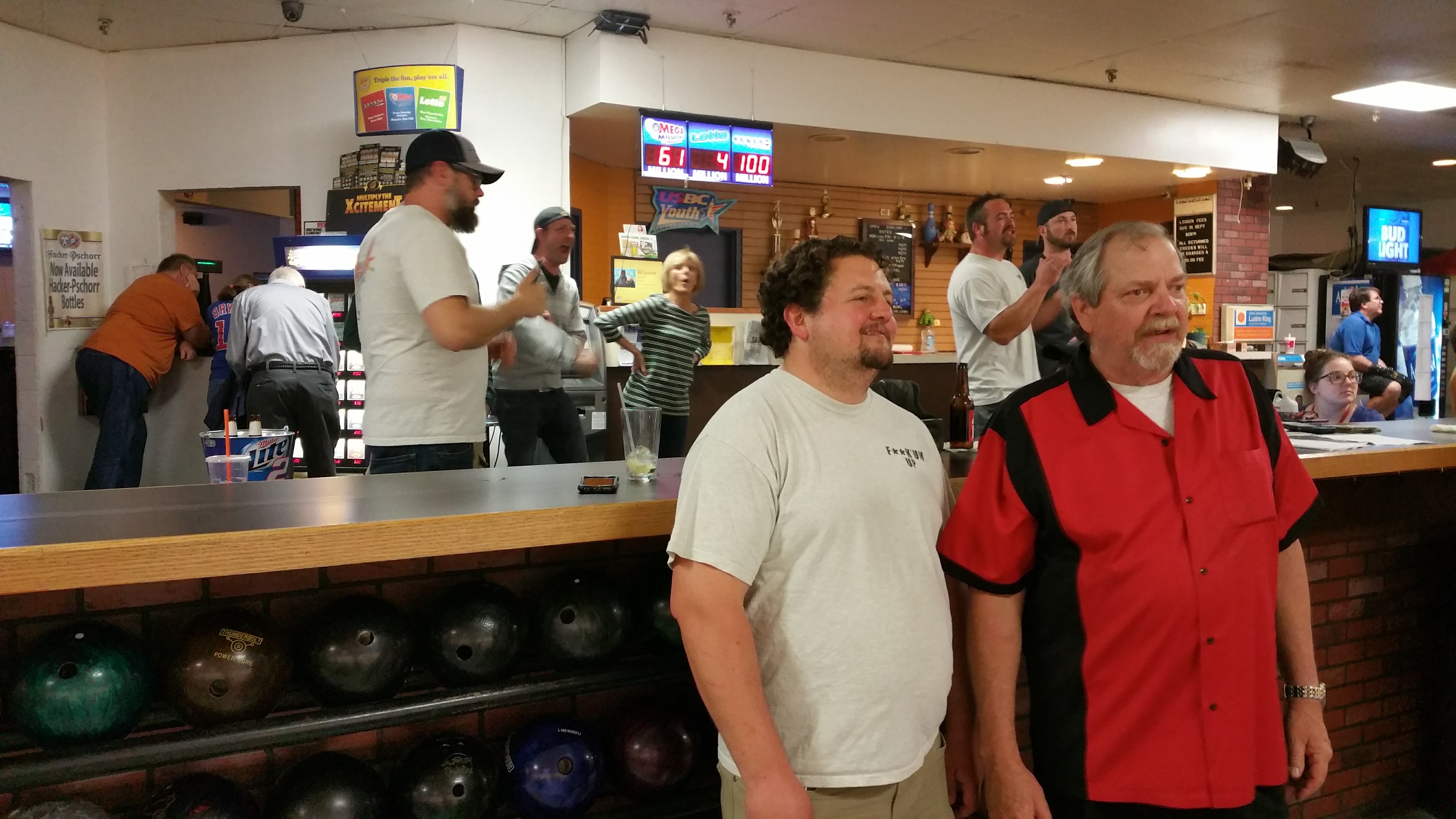 A candid photo of several adults socializing in a bowling alley, with bowling balls in the foreground and digital scoreboards in the background.