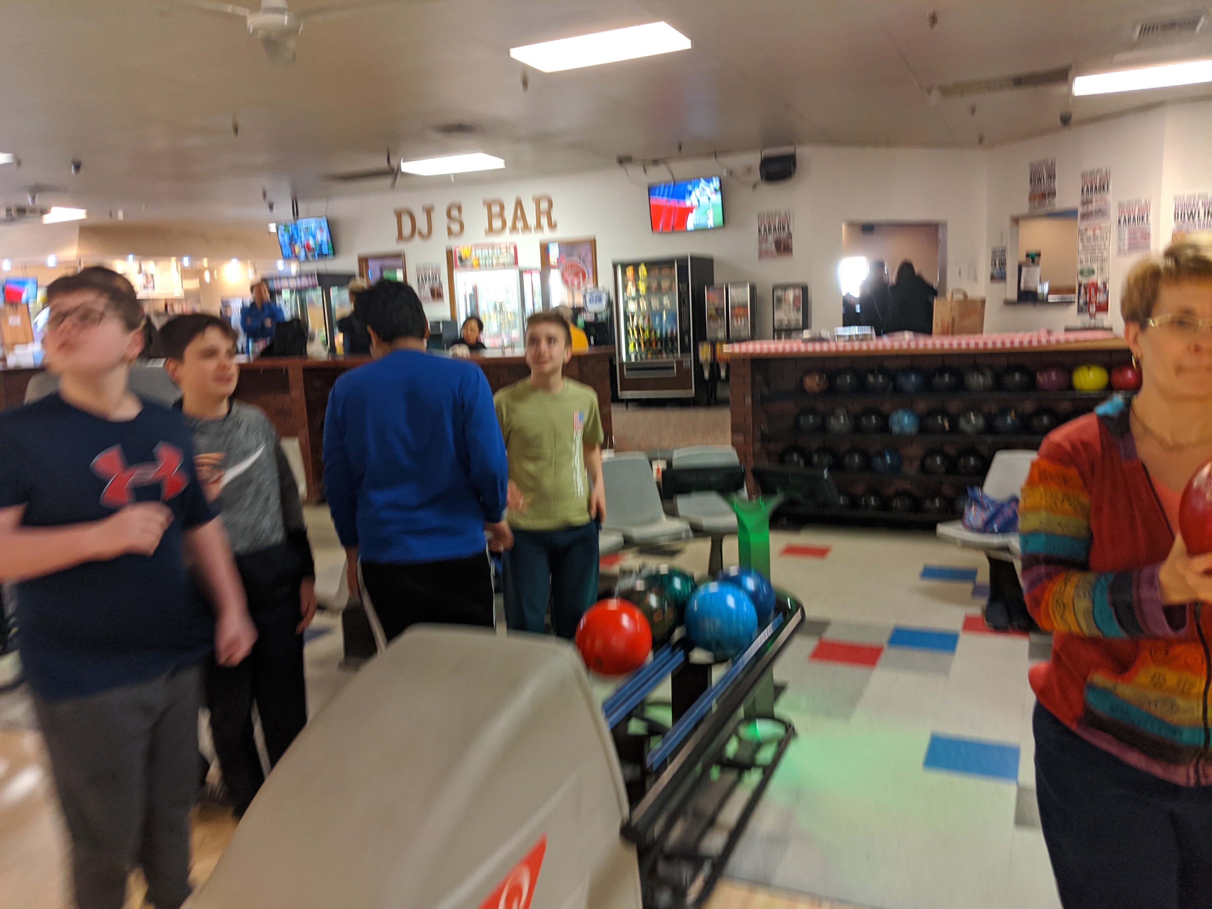 People bowling at a lively bowling alley, with colorful bowling balls on the return and a bar area visible in the background.