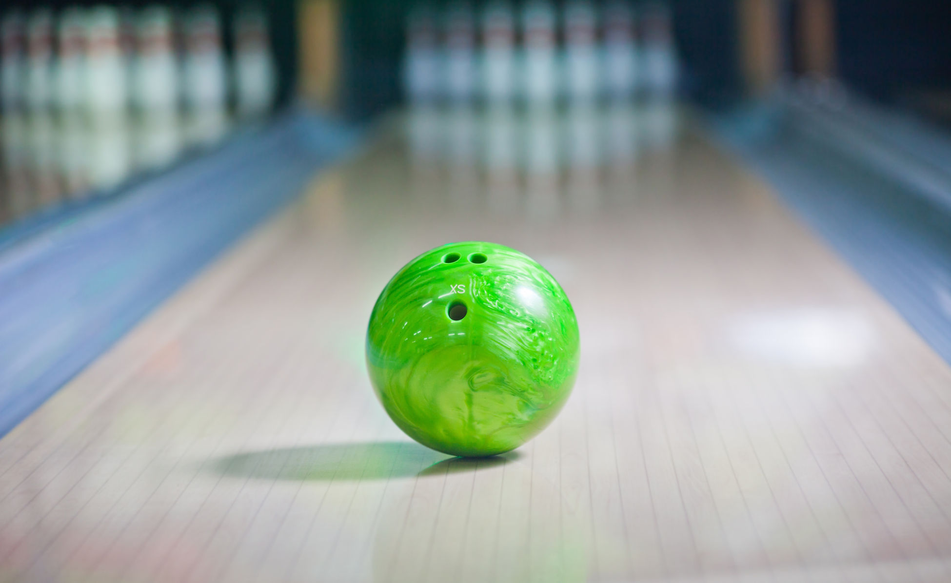 A bright green bowling ball with an 'XS' label rests on a polished wooden bowling lane, with pins blurred in the background.