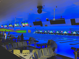 An indoor bowling alley lit with blue and purple cosmic bowling lights, showing multiple lanes, automatic scoring screens, and seating.
