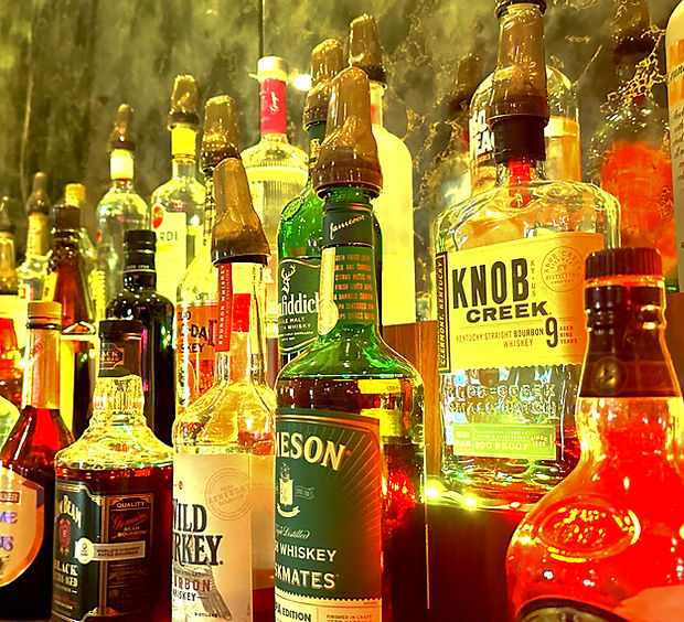 A close-up view of numerous liquor bottles with pour spouts, including Jameson and Knob Creek, illuminated by warm light.