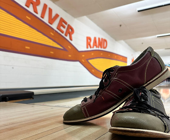Two pairs of well-worn bowling shoes rest on a wooden bowling lane, with the River Rand logo visible in the background.