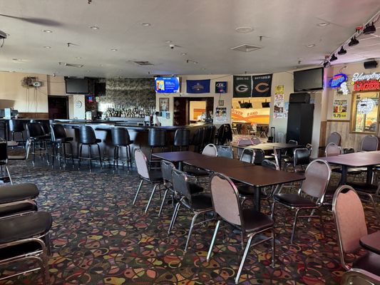 An empty bar and lounge area with dark wood bar stools, tables, and a colorful patterned carpet.