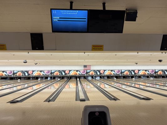 View down multiple bowling lanes with an automatic scoring screen visible above, showing a game for Howard and Ethel.