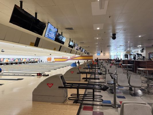 A wide view of a bowling alley with multiple lanes, overhead scoring monitors, and a bar and seating area.