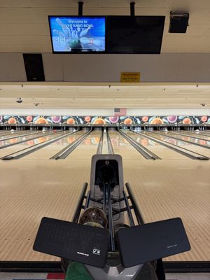 View of bowling lanes with a scoring screen displaying "Welcome to River Rand Bowl" and a bowling ball in the return.