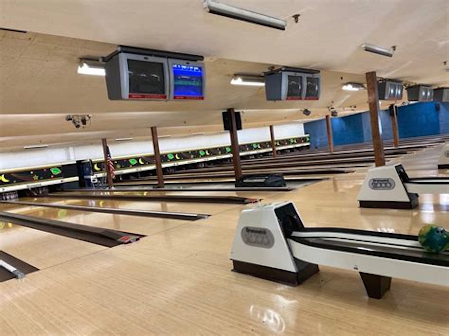 An interior view of a bowling alley with multiple lanes, overhead scoring monitors, and a bowling ball on a return.