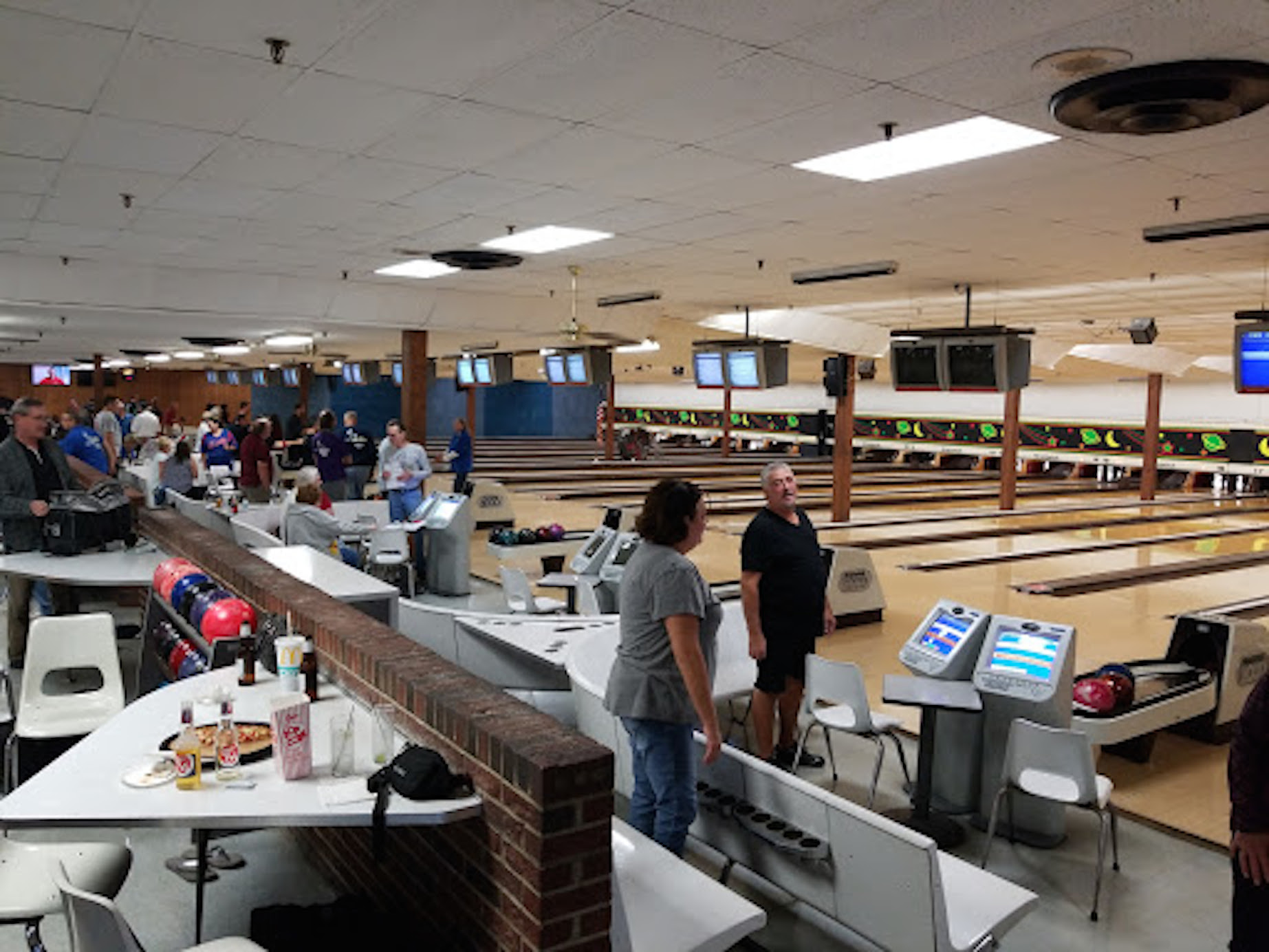 A busy bowling alley scene with multiple lanes, people bowling, automatic scoring screens, and food on tables.