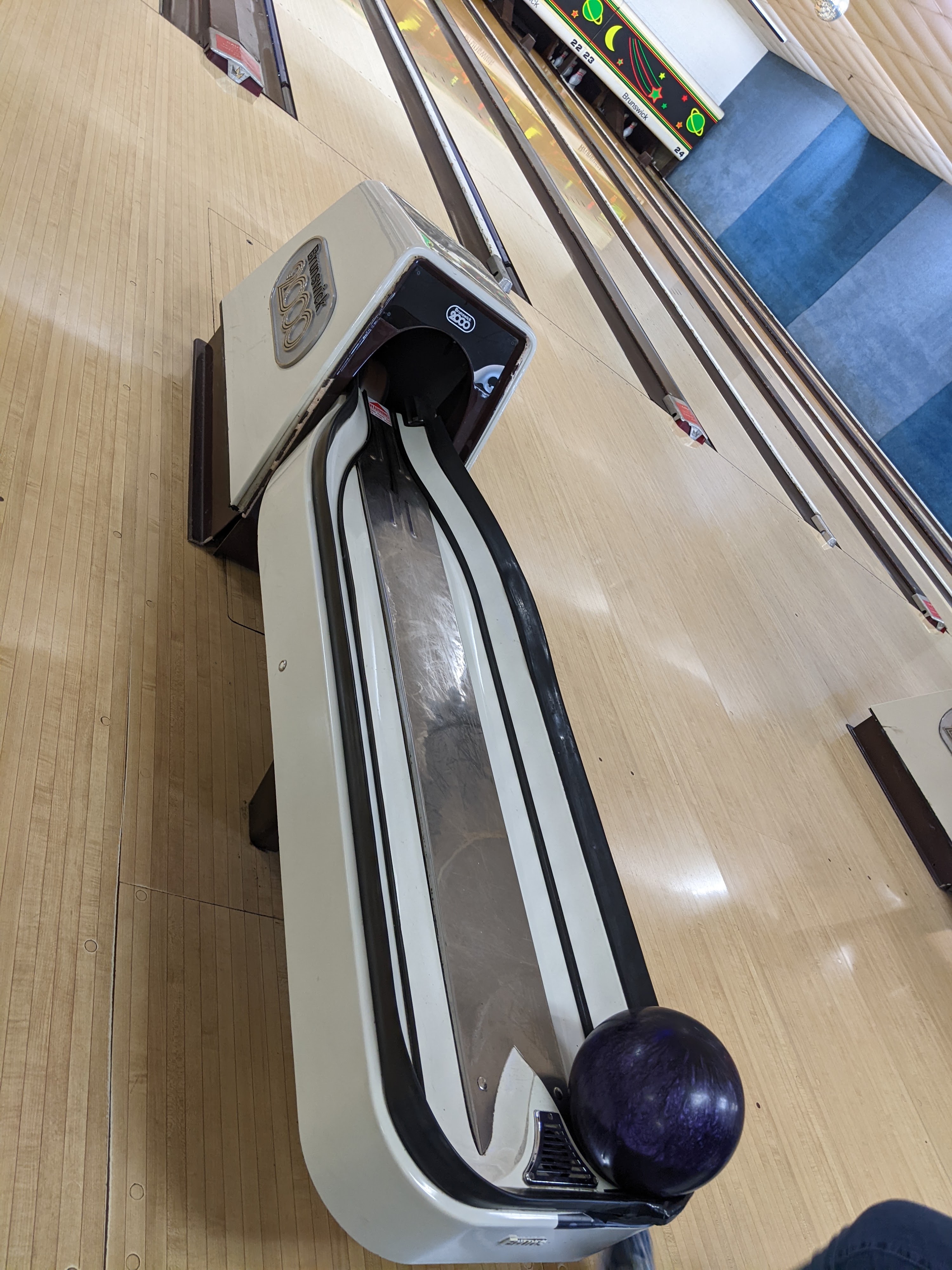 A close-up, high-angle view of a bowling lane with a purple bowling ball in the ball return.