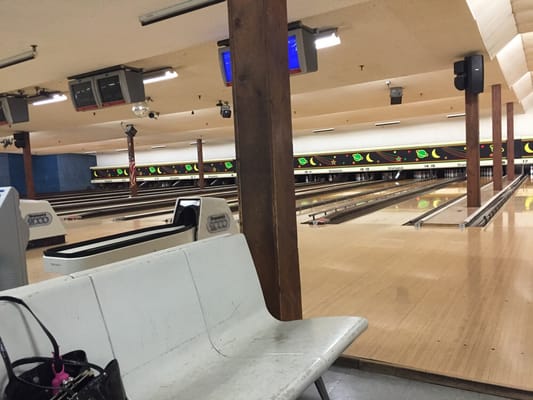 An empty bowling alley with multiple lanes, overhead automatic scoring screens, and a white bench in the foreground.