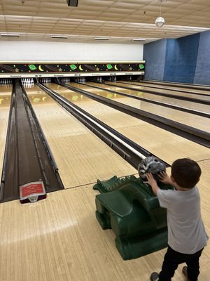 A young child from behind, using a green dinosaur bowling ramp to send a bowling ball down a lane.