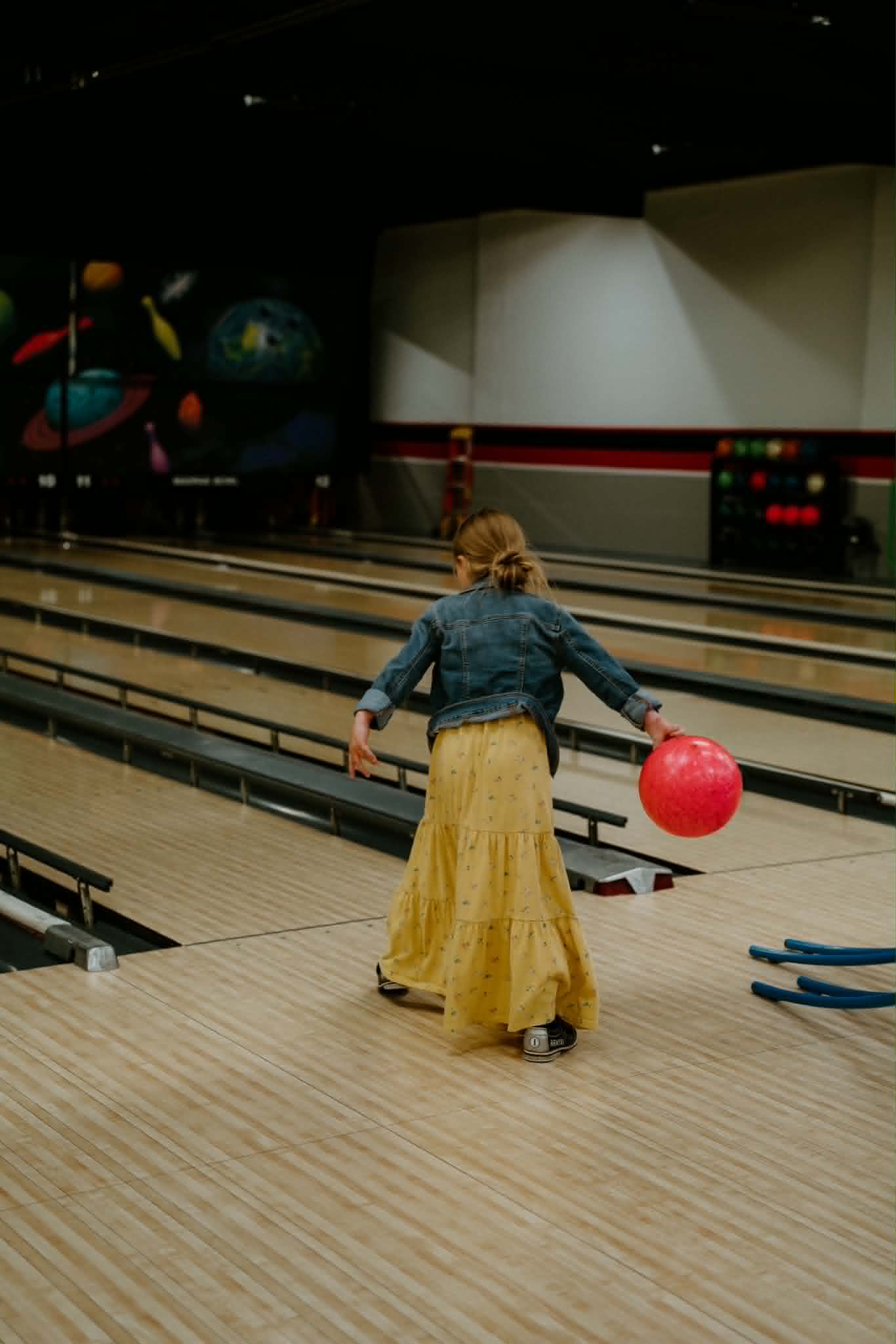 A young girl in a yellow dress and denim jacket prepares to bowl a red ball on a lane with bumpers.