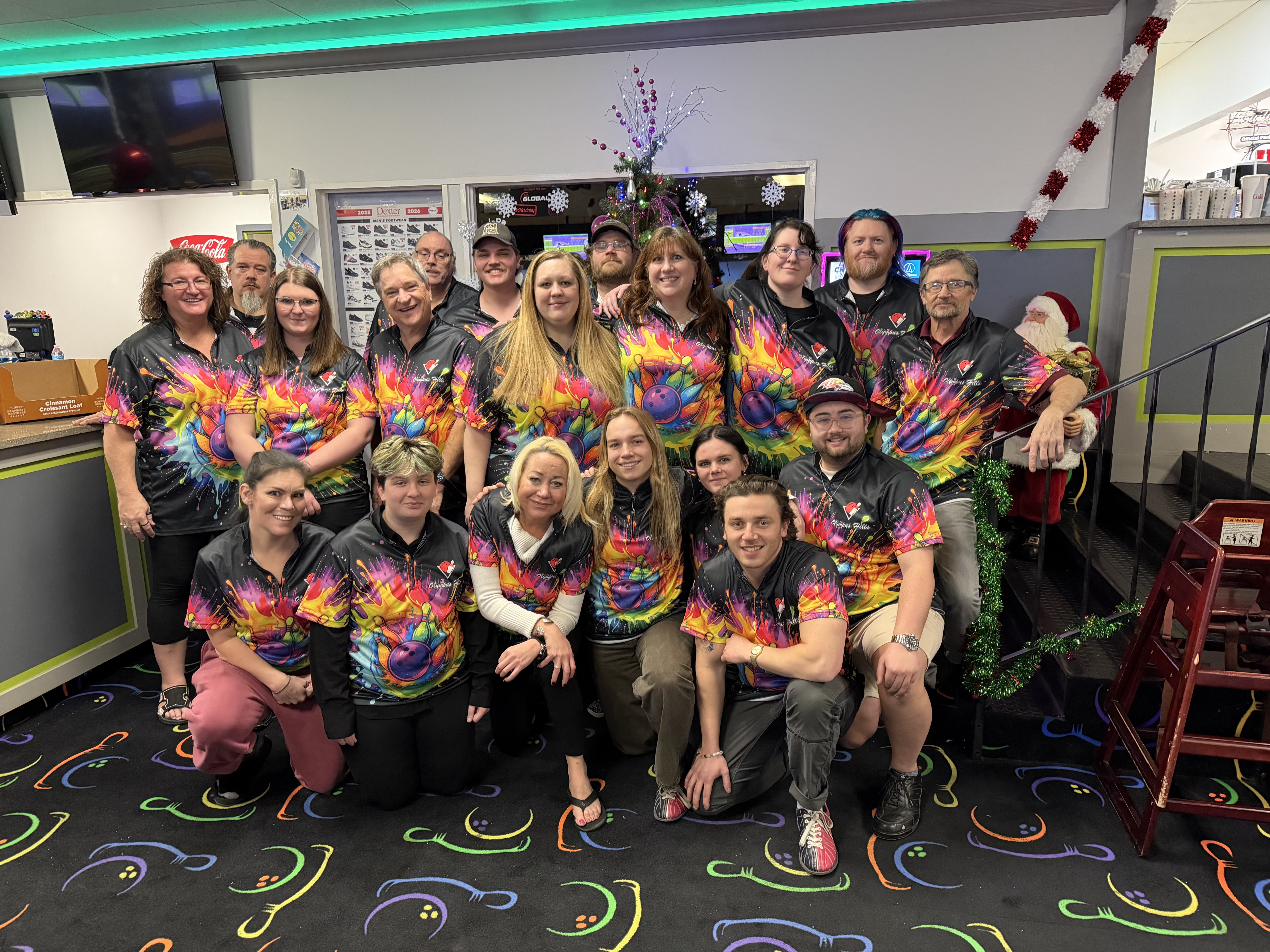 A large group of people in matching colorful bowling jerseys posing together in a bowling center with holiday decor.