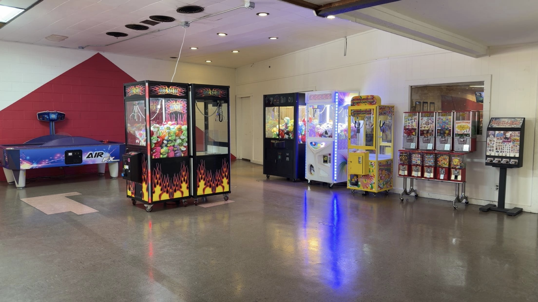 A clean, brightly lit arcade area with crane games, air hockey, and gumball machines on a polished floor.