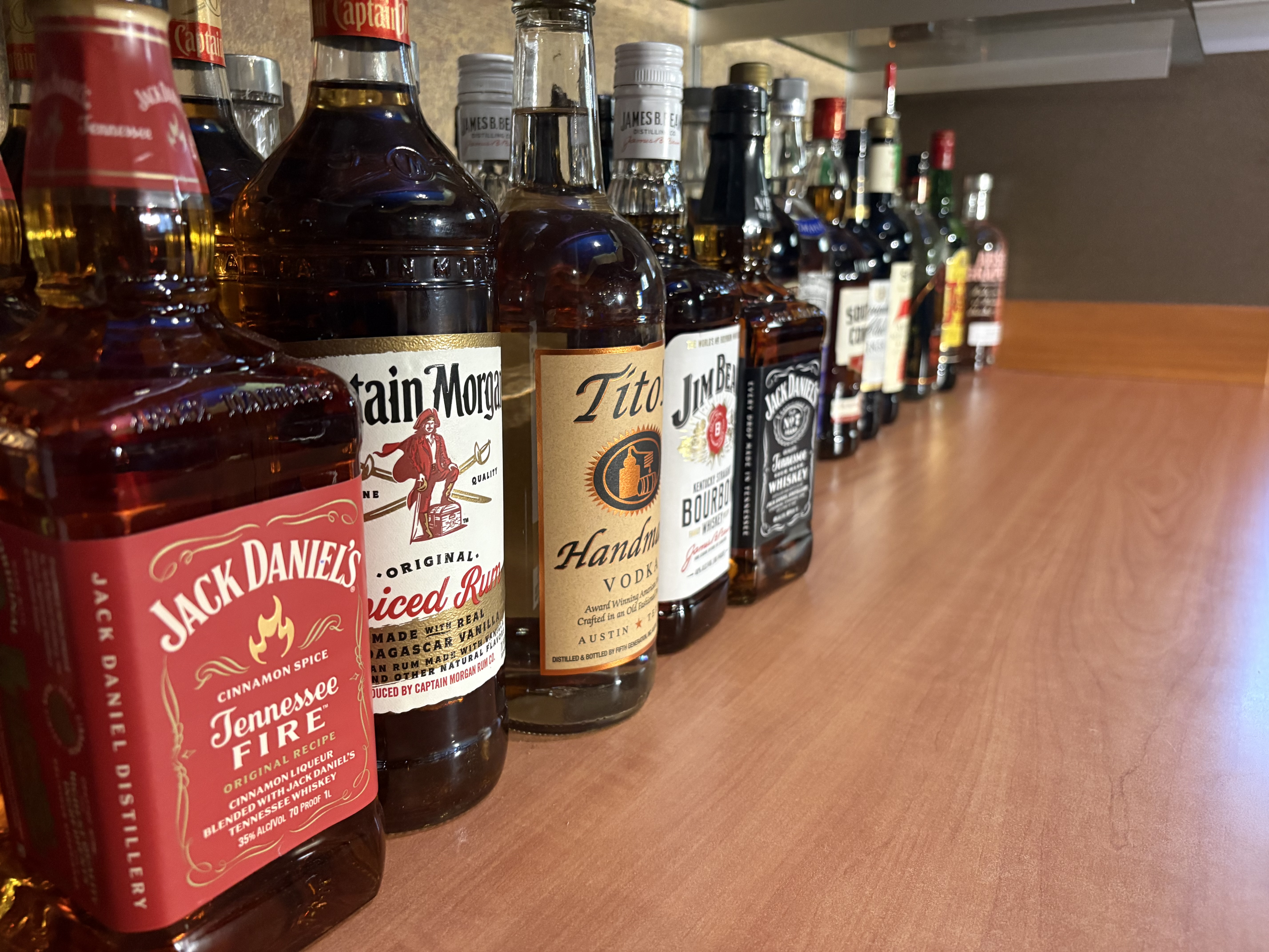 A close-up view of several liquor bottles, including Jack Daniel's, Captain Morgan, Tito's, and Jim Beam, lined up on a wooden bar.