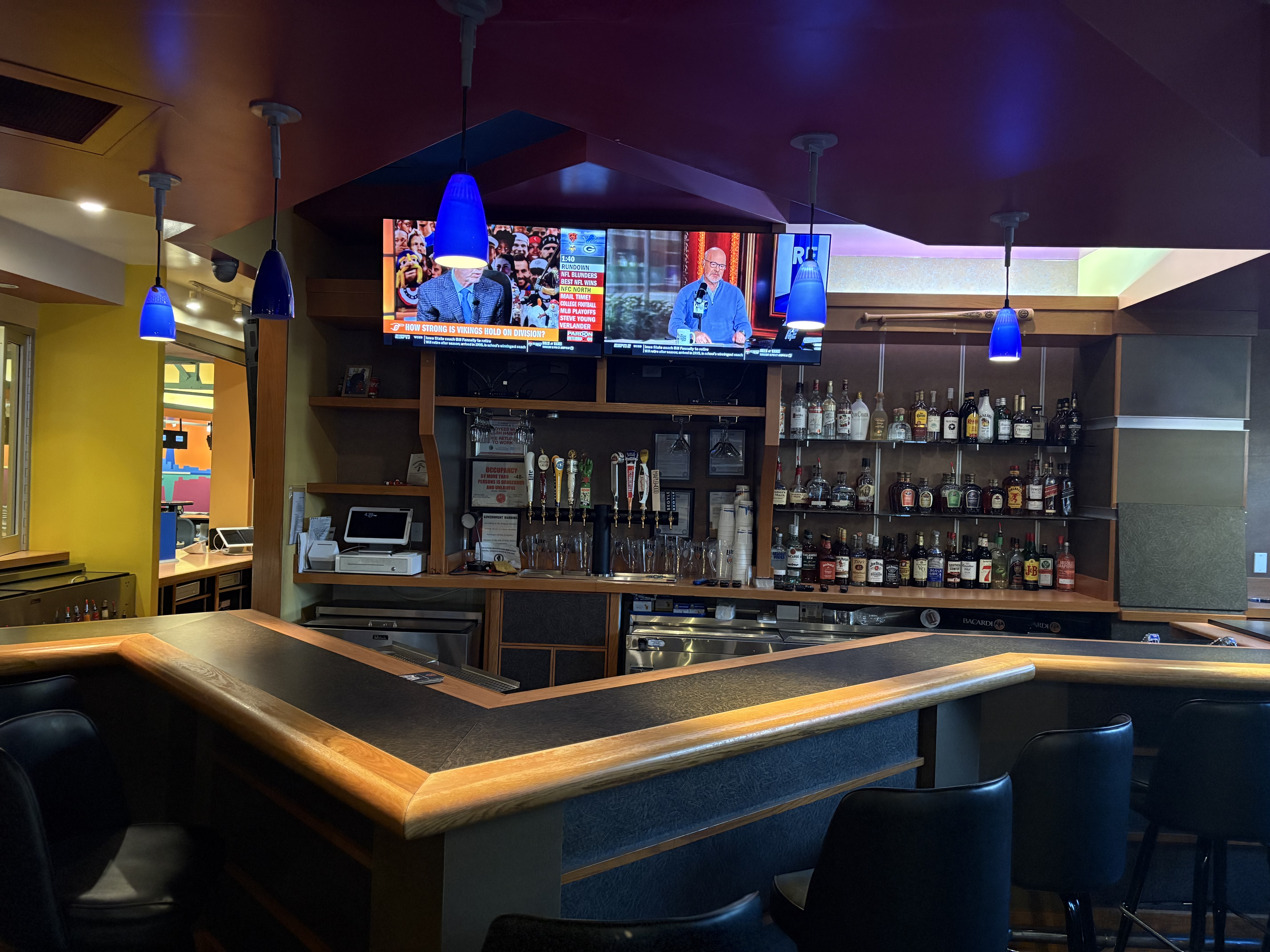 A well-stocked bar area with blue pendant lights, bar stools, and two televisions displaying sports.