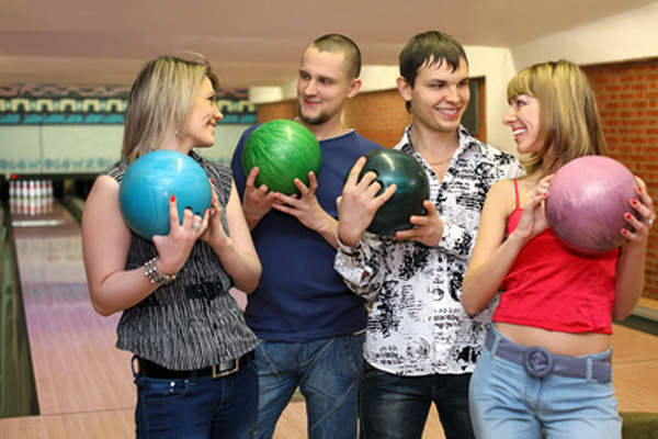 Four friends smiling while holding colorful bowling balls at a bowling alley.