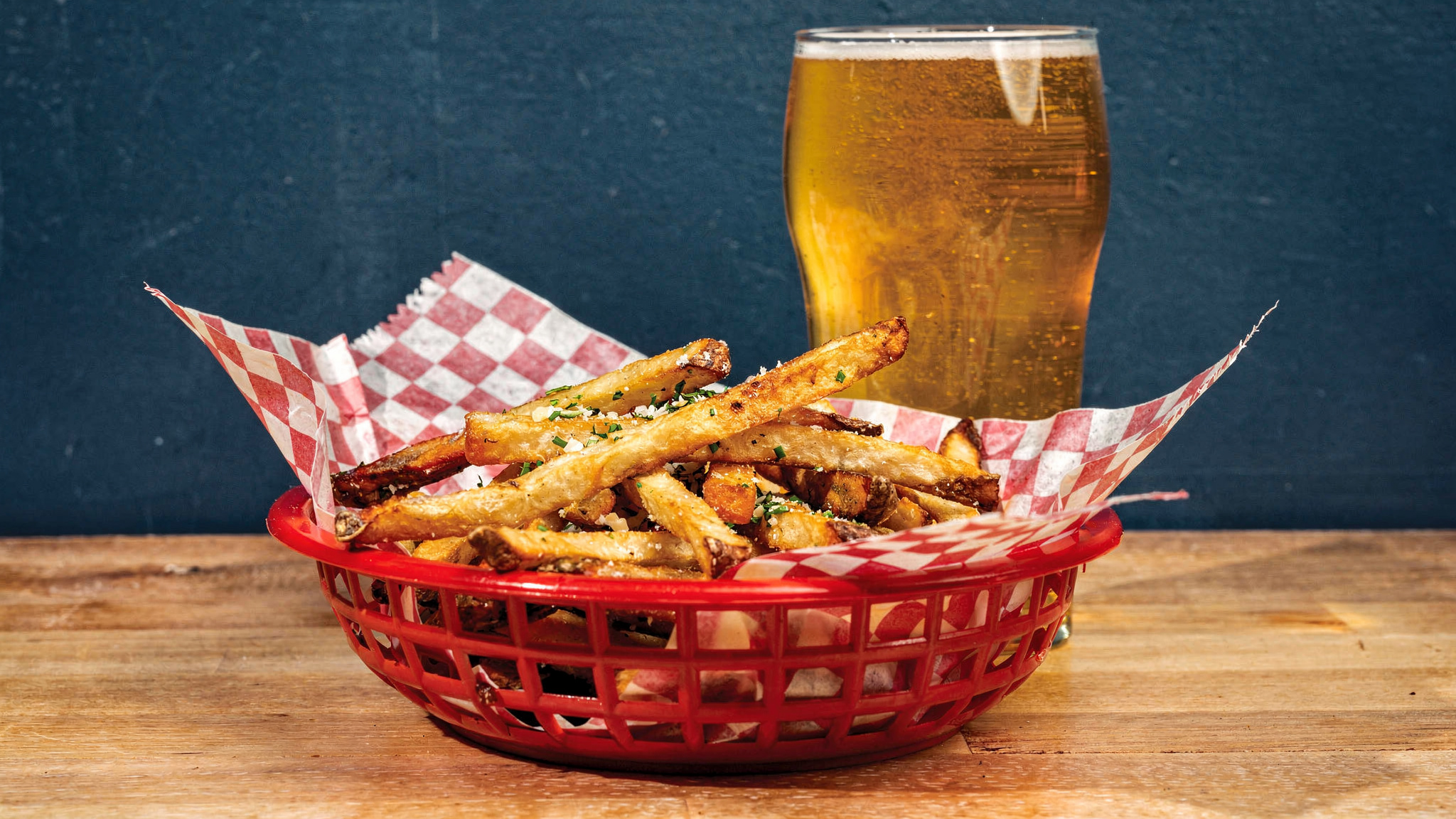 Seasoned french fries in a red basket next to a pint of beer on a wooden table.