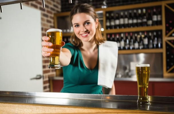 A smiling bartender handing a glass of cold beer across a bar counter.